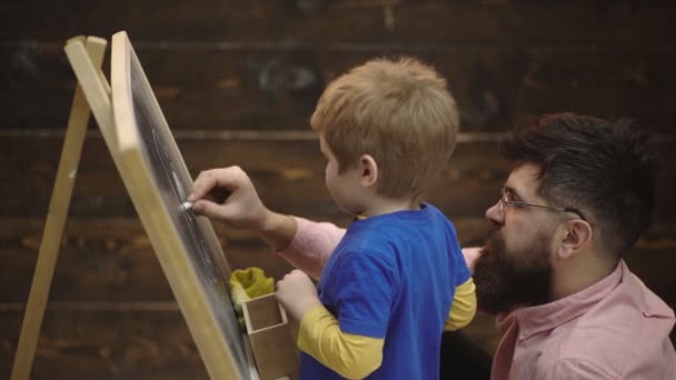 Крупним планом хлопчика і бородатого чоловіка, який пише крейдою на дошці. Студент і вчитель збоку перед дошкою. Батько або репетитор допомагають маленькій дитині малювати на фоні дерев'яних панелей . — стокове відео
