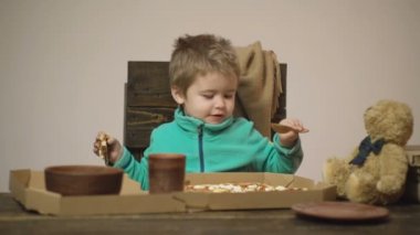 Arka plan. Lezzetli pizza. Küçük bir çocuk bir dilim pizza yiyor. Aç çocuk pizzadan bir ısırık alıyor. Beslenme kavramı. Yeşil kazak giyen çocuk tahta bir sandalyede otururken pizza yer..