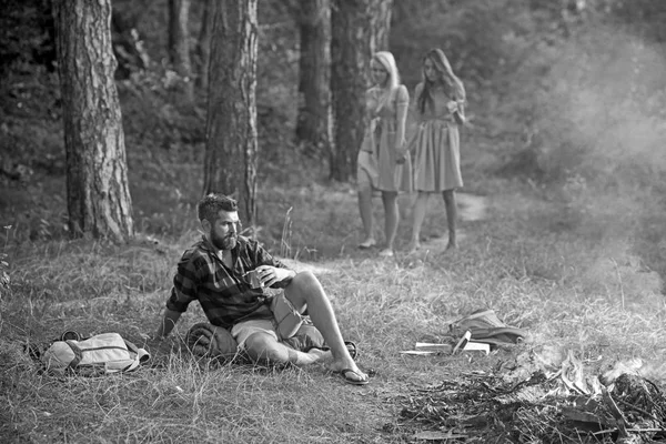 Piknik a napsütötte réten. Két lány vándor az erdőben, míg a gondolkodó ember ül a füvön. Brutális szakállas ember iszik kávét vagy teát, tábortűz. Barátság és a természet fogalmak — Stock Fotó