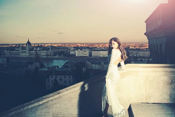Femme en robe de mariée blanche sur vue sur la ville du soir, la mode. Femme sensuelle avec les cheveux longs sur le balcon, beauté. Mariée maquillée. Fille avec look glamour dans le mariage — Photo