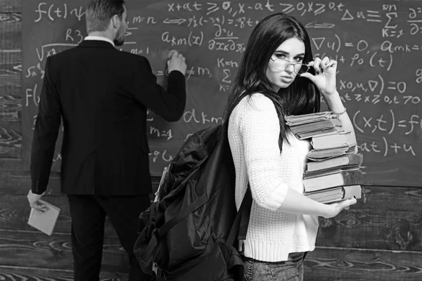 Sexy estudiante morena sosteniendo sus gafas mientras lleva un montón de libros. Joven chica universitaria en cárdigan rosa caminando delante de la pizarra verde — Foto de Stock