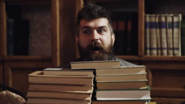 Man, nerd on surprised face between piles of books in library, bookshelves on background. Teacher or student with beard wears eyeglasses, sits at table with books, defocused. Nerd concept. — Stock Video