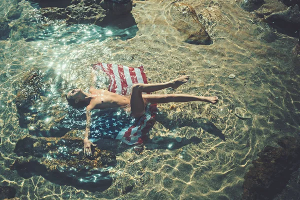 Καλοκαιρινές διακοπές και παραδείσου ταξίδι διακοπές. Ζευγάρι στην αγάπη με το προκλητικό σώμα, χαλαρώστε στην παραλία. Αγαπούν τις σχέσεις του γυμνό ζευγάρι στο νερό της θάλασσας. Αμερικανική σημαία σε νερό με σέξι γυναίκα και άνδρα έχουν παιχνίδια σεξ. — Φωτογραφία Αρχείου