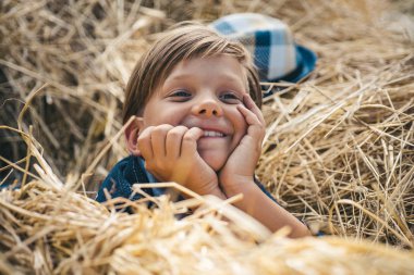 Başı samanlıkta şapkası olan neşeli bir adam. Çocuk sonbahar için çocuk kıyafetleri reklamı. Mutlu çocukluk. Köyde bir sonbahar tatilinde sevimli çocuk.