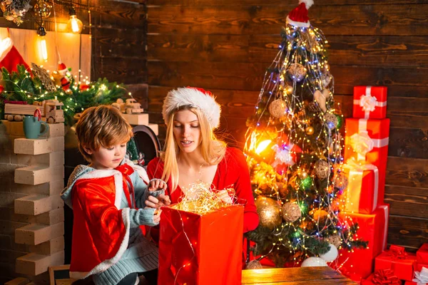 Young mother with Santa hat and the little boy are looking inside the box with Christmas light. Christmas tree background. Happy family. — ストック写真