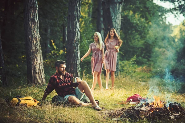 Hipster trinken Tee oder Kaffee am Lagerfeuer mit Mädchen auf verschwommenem Hintergrund. Hipster entspannen mit Becher am Lagerfeuer — Stockfoto