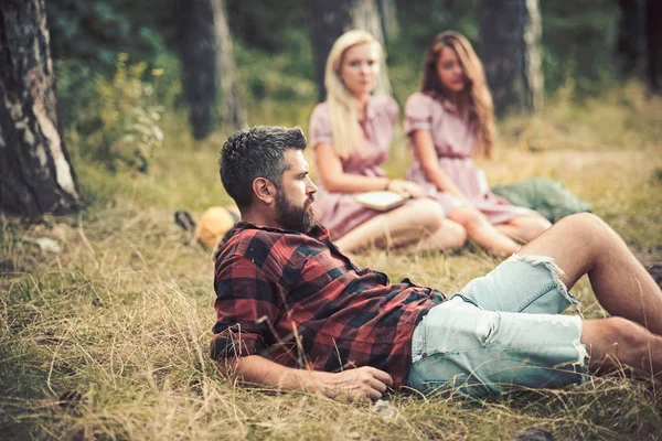 Um tipo barbudo deitado na relva no prado da floresta. Meninas sentadas ao lado da fogueira. Amigos acampar no final do verão. Relaxamento na floresta — Fotografia de Stock