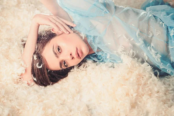 Woman with long hair in tender pajama relaxing. Girl on calm face lay on bed covered with feathers and fluff. Lady in transparent blue nightie lay on bed in heap of feathers. Airiness concept. — Stock Photo, Image