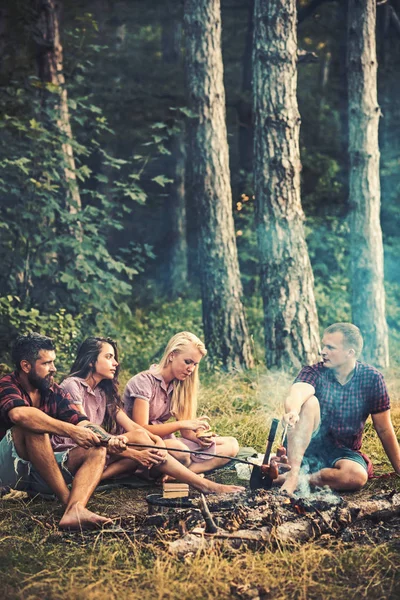 Friends talking around campfire. Men in lumberjack shirts cooking sausages outdoors. Two couples camping in woods