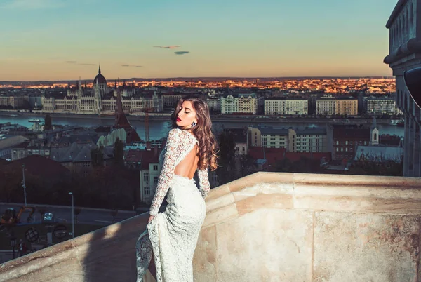 Femme sensuelle avec les cheveux longs sur le balcon, beauté. Femme en robe de mariée blanche sur vue sur la ville du soir, la mode. Mariée maquillée. Style de modèle de mode et coiffure. Fille avec look glamour . — Photo