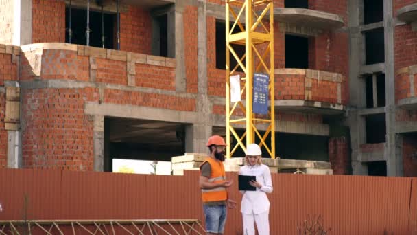 Gli ingegneri discutono la costruzione dell'edificio. Giovane ingegnere barbuto moderno o architetto con costruttore femminile sullo sfondo della costruzione di edifici. Due caporeparto discussione sul cantiere . — Video Stock