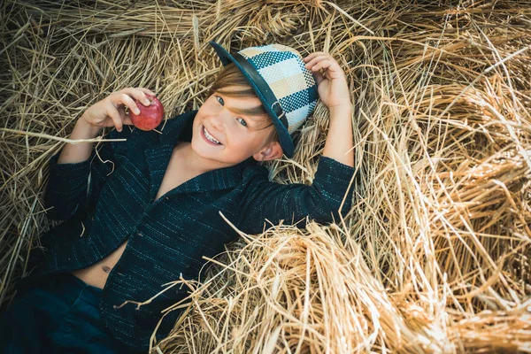 Fair-haired boy lies on hay background and eats an apple. Sale for entire autumn collection, incredible discounts. Smiling boy holds bitten apple with one hand. Back to school. Bye summer - hi autumn.