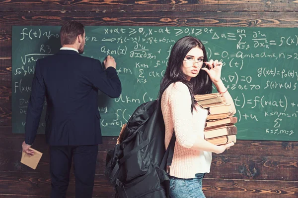 Brunette student met stapel boeken permanent in klas terwijl hoogleraar is het schrijven van formule op groen bord — Stockfoto