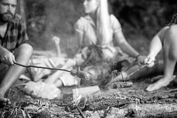 Cocinar salchichas en palos sobre llamas de fogata, acampar durante la noche en verano. Grupo de amigos sentados junto a la hoguera — Foto de Stock