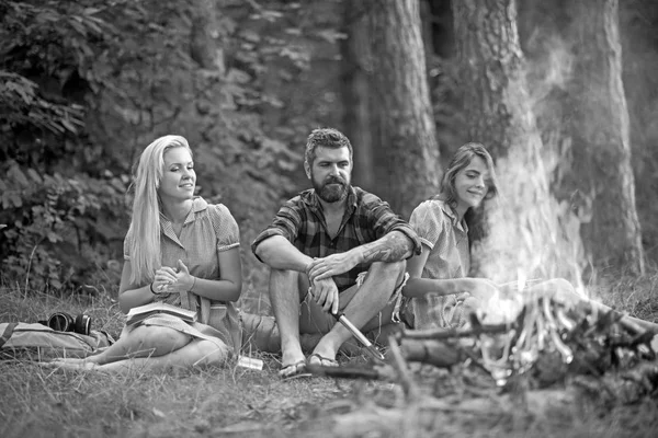 Lächelnde Freunde genießen die letzten Sommertage im Freien. Jugendliche sitzen am Lagerfeuer. Mädchen lesen Bücher, während sie auf Gras sitzen — Stockfoto