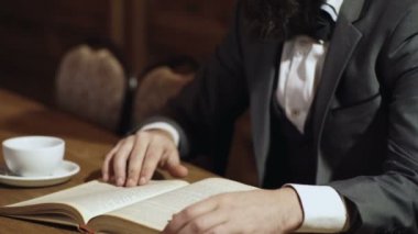 Açık bir kâğıt kitap tutan, sayfaları parmaklarıyla çeviren ve çay içen bir adamın yakın çekimi. Mans el bir sayfa dönüm. Mans el çevirme kitap sayfası. Kütüphanede Aristokrat.