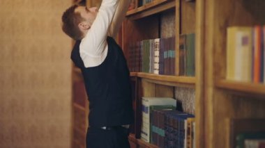 Yelekgiyen adam kütüphanedeki kitaplığından bir kitap seçer. Ciddi yüzlü olgun bir adam. Profesör büyük kütüphanede duruyor ve kitap seçin. Çalışma, öğrenme, öğretmek, tarih, edebiyat kavramı.