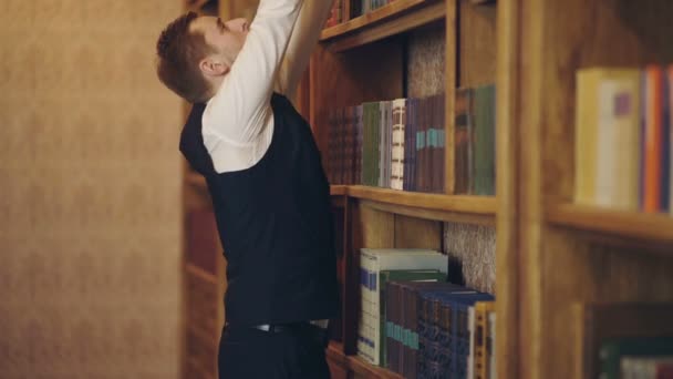 L'homme en gilet choisit un livre dans une bibliothèque. Homme mûr avec un visage sérieux. Professeur se tient dans la grande bibliothèque et choisir le livre. Étude, apprentissage, enseignement, histoire, littérature concept . — Video