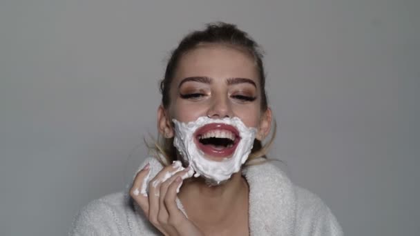 Soin de la peau et concept de rasage. Femme visage recouvert de mousse. Lady se soucie de la peau lisse. Fille avec le visage souriant en peignoir couvrant son visage de mousse pour le rasage, fond gris . — Video