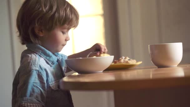 Retrato de un hermoso niño desayunando en casa. El niño en la cocina en la mesa comiendo. Riéndose lindo niño bebé niño sentado en trona y comer en fondo de cocina . — Vídeos de Stock