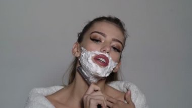 Bayan yüzü usturanın keskin bıçak ile tıraş. Kadın yüzü köpük ile kaplı olan ustura elinde tutar. Kız meşgul yüzündeki bornoz, gri arka plan giyer. Kuaför ve tıraş kavramı.