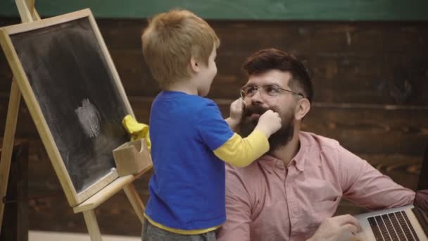 Kid pulling beard of the guy. Boy is having fun with his teacher in classroom. Charming man fulfills the boys desire. Smiling boy playing with beard of man next to him. Individual education concept. — Stock Video