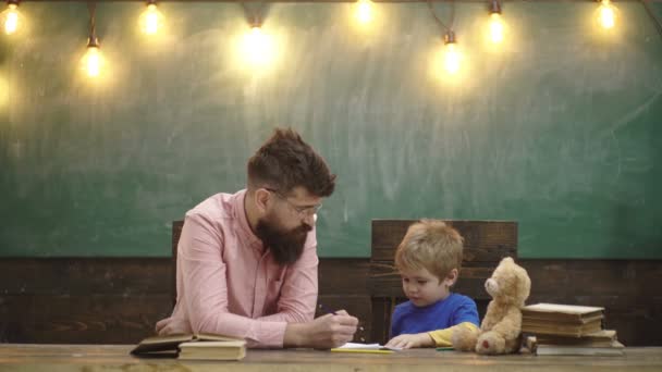 Lehrer mit stylischem Schnurrbart und Bart und kleinem Kind, das Buchstaben ins Lesebuch schreibt. Netter Junge zeichnet ein Bild mit bunten Bleistiften. Kunststunde im Kindergarten. Vater lehrt seinen Sohn zeichnen. — Stockvideo