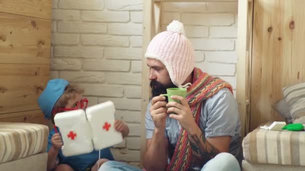 Sick man in a cap and a scarf drinks tea, and a boy in a medical uniform gives him medicine from a first-aid kit. Father and kid playing clinic. Boy dressed as doctor playing with dad. — Stock Video