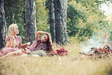 Doğal makyajlı kadınlar yeşil çimlerde rahatlar. Şenlik ateşinde uzun saçlı güzel kızlar. Ormanda eski moda elbiseli moda kadınları. Yaz tatili kavramı. Kamp gezisi, kitap okumak, klasik.