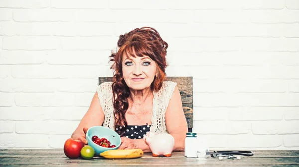 Senior vrouw met spaarvarken, fruit, geneeskunde, stethoscoop aan tafel — Stockfoto