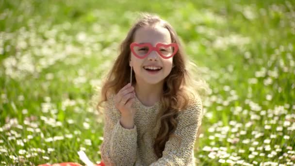 Flickan sitter på gräs på grassplot, grön bakgrund. Barnflickan tillbringar fritid utomhus. Bästa semesterkoncept. Barn som poserar med Crown och glasögon Photo Booth attribut. Rolig och lycklig barndom. — Stockvideo