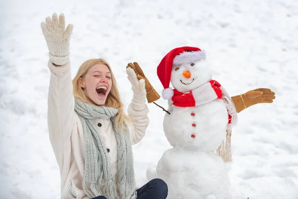 Vzrušená dívka s sněžným mužem na zasněžené zimní procházce. Dělat sněti a zimní zábavu. Roztomilý sněhobák v klobouku a skalf na zasněženém poli. — Stock fotografie
