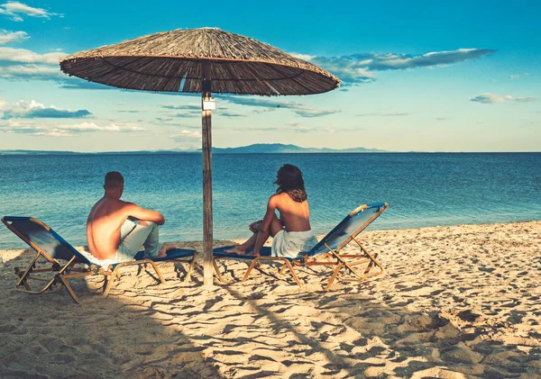 Pareja joven sentada en una arena y mirando a un mar bajo el paraguas . — Foto de Stock