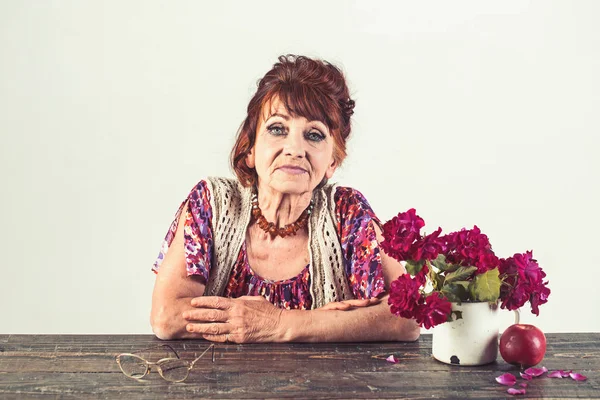 Senhora idosa posar com flores, maçã, óculos na mesa . — Fotografia de Stock