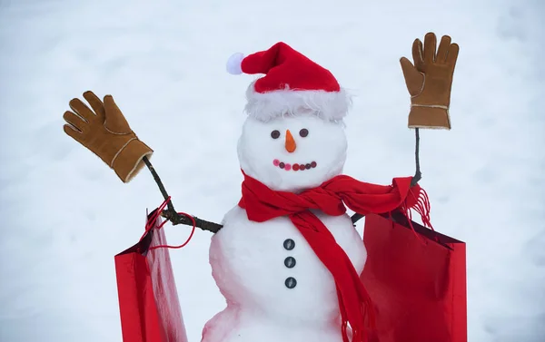 雪人戴围巾，戴帽子，带购物袋。雪中快乐有趣的雪人。白色雪背景的圣诞雪人。快乐的雪人与礼品盒站在冬季圣诞景观. — 图库照片
