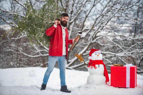 Bärtiger Mann mit Schneemann trägt Weihnachtsbaum im Wald. Ein gutaussehender junger Mann mit Schneemann trägt einen Weihnachtsbaum. Schneemann mit Geschenk. — Stockfoto
