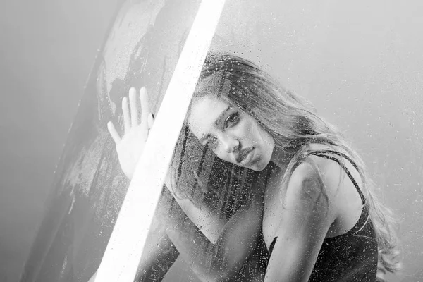 Shower and hygiene spa treatment. Sexy woman behind plastic sheet with water drops. Fashion and beauty. Rain drops on window glass with face of girl. Window with water drops before girl with makeup
