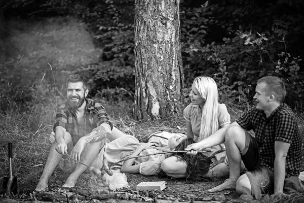 Happy men and women roast sausages on fire. Friends have picnic at bonfire in wood. Boyfriends and girlfriends enjoy camping food. People relax on summer nature. Camping and cooking food on vacation