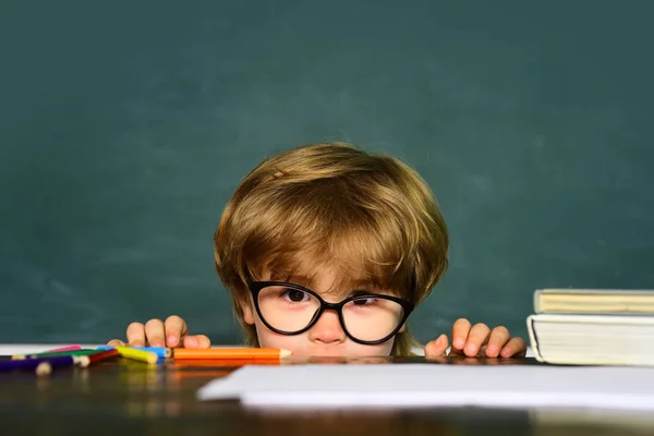 Rapaz bonito com expressão facial feliz perto da secretária com material escolar. Kid está aprendendo na aula no fundo do quadro negro . — Fotografia de Stock