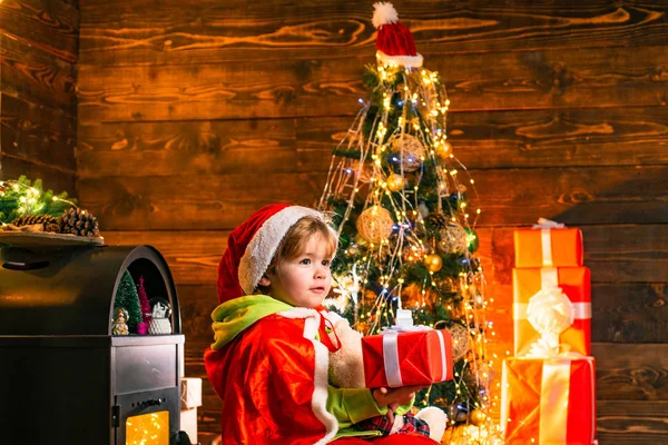 Curios menino está brincando com brinquedos junto à árvore de Natal. O miúdo está a usar roupa de Pai Natal. Conceito de férias. — Fotografia de Stock