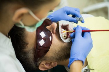 Gülümseyen sevimli bir kamera. Modern diş kliniğinde kadın dişçi hasta dişlerini aynayla kontrol ediyor. Genç adamın arka plandaki bulanık ışıkta diş hekimi incelemesi.