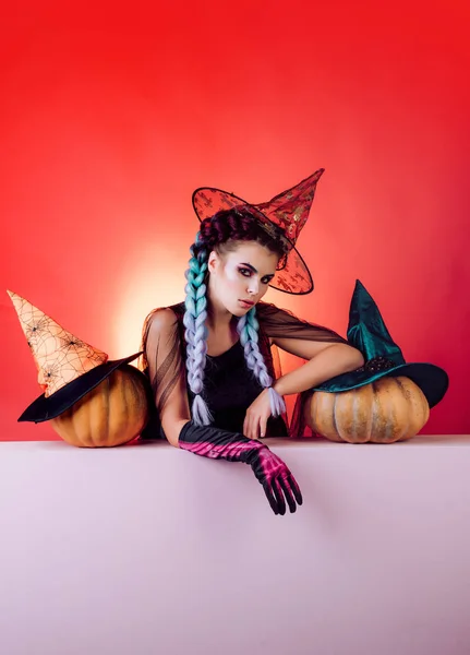 Mujer joven gótica feliz en traje de Halloween bruja con sombrero de pie fondo rojo. Hermosa mujer sorprendida con sombrero de brujas y traje que sostiene la calabaza. Vacaciones, decoración y concepto de fiesta . — Foto de Stock