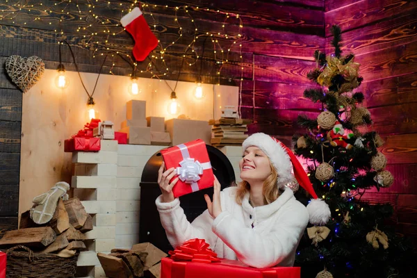 Sexy Santa Clause woman in elegant dress. Christmas and new year concept. Woman in red Santa hat holding present. Blonde Santa woman holding gift box at vintage wall. Christmas fashion.
