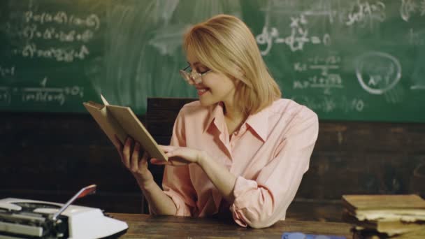 Lachende jonge vrouw in een roze overhemd is het lezen van een boek in de buurt van een groen schoolbord met formules erop geschreven. Jonge mooie blonde meisje student vóór de les. Docent in klaslokaal. Docent en student. — Stockvideo