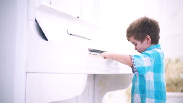 Liten pojke tycker om att spela piano, första gången. Barn stativ nära piano keyboard, vit bakgrund. Kid tillbringa fritid nära musikinstrument. Pojke söt och bedårande sätter fingret på tangentbordet av piano. — Stockvideo