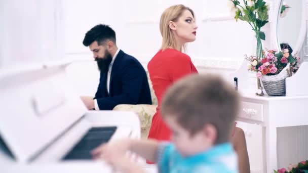 Niño toca el piano, su madre hace maquillaje delante del espejo y su padre lee un libro en un costoso interior blanco, foco de transferencia. Los padres ricos disfrutan de la paternidad. Niño adorable tocar el piano . — Vídeos de Stock