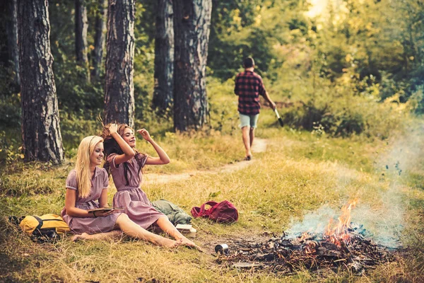 Mulheres felizes com o homem desfocado na floresta de verão. As mulheres de moda em vestidos retro relaxam na fogueira. Meninas de beleza com cabelo longo sorriso na grama verde. Férias camping viagens e livros de leitura, vintage — Fotografia de Stock
