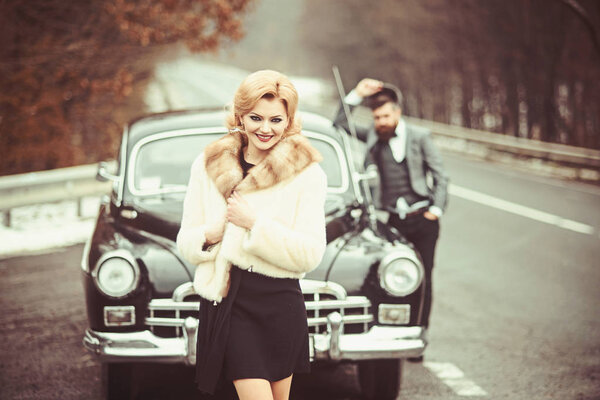 Retro collection car and auto repair by mechanic driver. Bearded man and sexy woman in fur coat. Couple in love on romantic date. Travel and business trip or hitch hiking. Escort of girl by security.