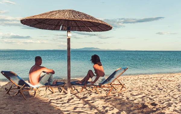 Pareja joven sentada en una playa de arena tropical bajo el paraguas . — Foto de Stock