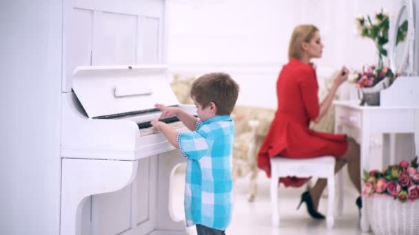 Anak berdiri di dekat keyboard piano, latar belakang interior putih. Konsep pendidikan musisi. Orang tua kaya menikmati keibuan. Boy adorable mencoba untuk bermain alat musik piano, sementara ibu melakukan makeup . — Stok Video